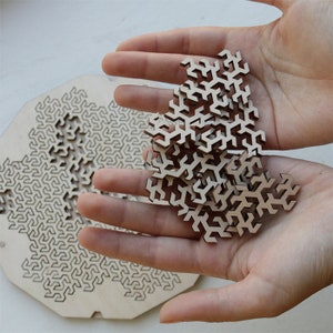 May include: A wooden puzzle with a geometric design. The puzzle pieces are in the shape of a snowflake and are scattered on a hand. The puzzle is made of light brown wood.