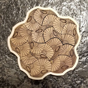 May include: A wooden coaster with a brown and black geometric design. The design features a repeating pattern of curved lines and circles.