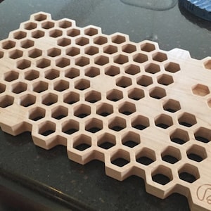 Puede incluir: Una tabla de cortar de madera con un diseño de panal de abeja. La tabla está hecha de madera clara y tiene un acabado natural. El diseño de panal de abeja está cortado en la tabla, creando un aspecto único y elegante.