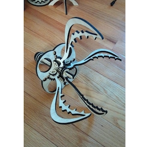 Könnte beinhalten: Ein mechanisches Modell aus Holz, lasergeschnitten, einer Kreatur mit mehreren beweglichen Armen und einem zentralen Getriebesystem. Das Modell besteht aus hellem Holz mit schwarzen Akzenten und ist teilweise montiert.