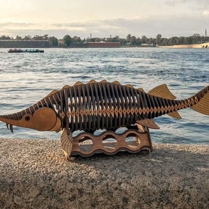 Könnte beinhalten: Ein Holzmodell eines Störs mit einem detaillierten, segmentierten Körper und einer langen, spitzen Schnauze. Das Modell ist auf einem Holzständer montiert und vor dem Hintergrund eines Flusses und einer Stadtkulisse ausgestellt.