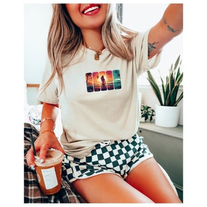 May include: A woman wearing a beige t-shirt with a graphic of a sunset and palm trees. She is wearing black and white checkered shorts and holding a cup of coffee.