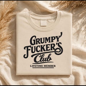 May include: A beige crew neck t-shirt featuring black text that reads Grumpy Fucker's Club in a stylized script font, with the words LIFETIME MEMBER printed in a smaller, bold, sans-serif font directly underneath the main graphic design.