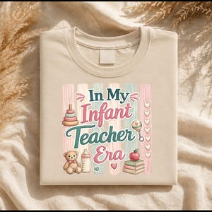 Könnte beinhalten: Ein beiges T-Shirt mit einem Grafikdruck und dem Text In My Infant Teacher Era. Das Design zeigt Illustrationen eines Stapelspielzeugs, eines Teddybären, einer Babyflasche, einer Rassel, eines Apfels und eines Bücherstapels vor vertikalen Streifen.