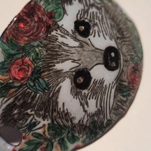 May include: Oval brooch with a hand-painted sloth design. The sloth has a grey and white face with black eyes. The brooch is surrounded by red roses, green leaves, and a blue border.