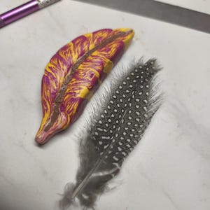 Peut inclure: Une plume en argile polymère faite à la main, aux couleurs jaune, violet et marron, à côté d'une plume naturelle avec des taches noires et blanches. La plume en argile a un aspect texturé et marbré. La plume naturelle est grise et blanche.