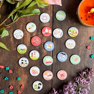 Puede incluir: Un conjunto de 20 fichas redondas de colores con diversas ilustraciones y texto. Las fichas representan actividades como jugar al aire libre, leer un libro, poner la mesa y sacar la basura. Las fichas están sobre una superficie de madera con una vela y flores.