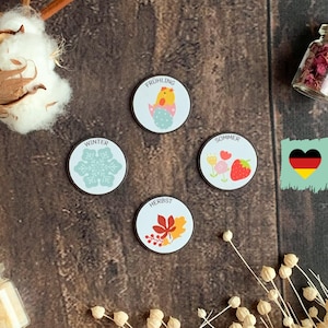 Può includere: Quattro magneti rotondi e colorati con parole tedesche per le stagioni: Frühling (Primavera), Sommer (Estate), Herbst (Autunno), Winter (Inverno). Ogni magnete presenta un'immagine stagionale: un pulcino, fiori, foglie e un fiocco di neve.