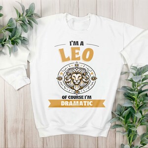 May include: White long-sleeved sweatshirt with a gold graphic of a lion and the text "I'm a Leo Of course I'm Dramatic".