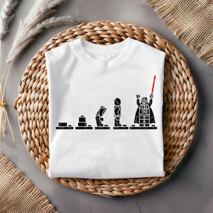 Puede incluir: Camiseta blanca con un diseño gráfico negro que representa la evolución de Darth Vader, al estilo Lego. El diseño muestra la progresión de bloques a una figura completa de Darth Vader con un sable de luz rojo. La camiseta está doblada sobre una cesta tejida.