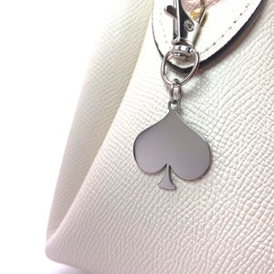 May include: A white leather handbag with a silver spade-shaped charm attached to a silver hook.
