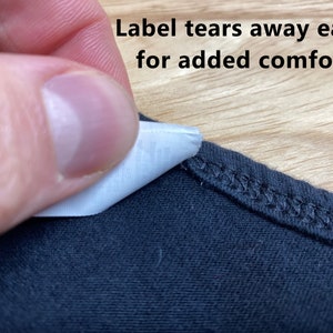 May include: A close-up of a black t-shirt with a white tag that says "Label tears away easily for added comfort!"