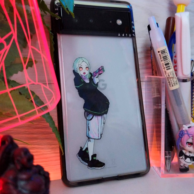 May include: A white phone case with a black border and a sticker of an anime character with green hair holding a gun. The character is wearing a black jacket with green accents and white trousers. The phone case has the Google logo on the back.