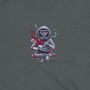 May include: A dark heather grey t-shirt featuring a cartoon illustration of an astronaut monkey meditating in space. The monkey is wearing a white spacesuit with red accents and is holding a cup of coffee. The illustration is surrounded by stars and planets.
