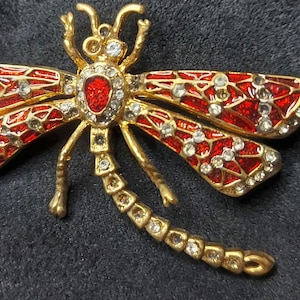 May include: A gold-toned dragonfly brooch with red enamel wings and a central red stone. The wings are detailed with clear crystals. The brooch has a detailed body and antennae, creating a sparkling, elegant accessory.