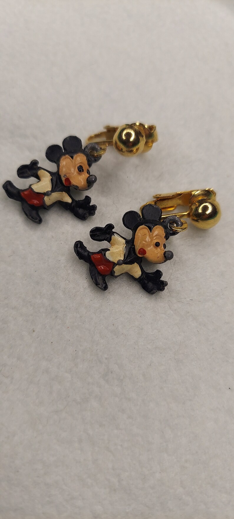 May include: A pair of vintage Mickey Mouse clip-on earrings. Each earring features a running Mickey Mouse figure in black, red, and tan. The clip-on mechanism is gold-toned.