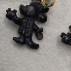 May include: Black clip-on earrings featuring a cartoon character design. The earrings have a gold-colored clip and a black figure with outstretched arms. The earrings are likely vintage or retro.