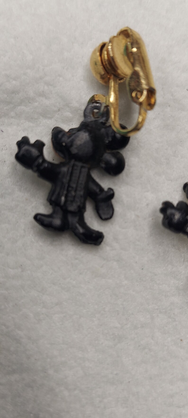 May include: Black clip-on earrings featuring a cartoon character design. The earrings have a gold-colored clip and a black figure with outstretched arms. The earrings are likely vintage or retro.