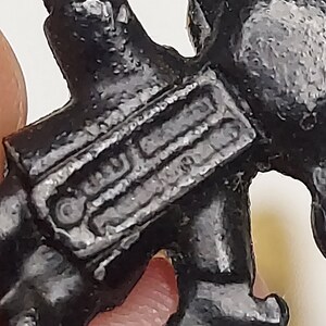 May include: Close-up of a small, black, stylized figurine. The figurine has a detailed, textured surface and appears to be a miniature representation of a person or character. The figure is held by a person's fingers.