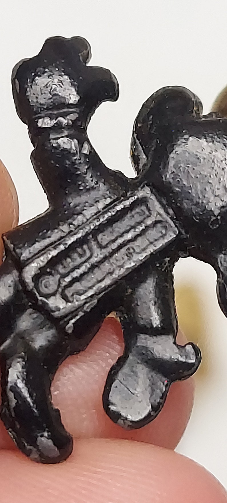 May include: Close-up of a small, black, stylized figurine. The figurine has a detailed, textured surface and appears to be a miniature representation of a person or character. The figure is held by a person's fingers.
