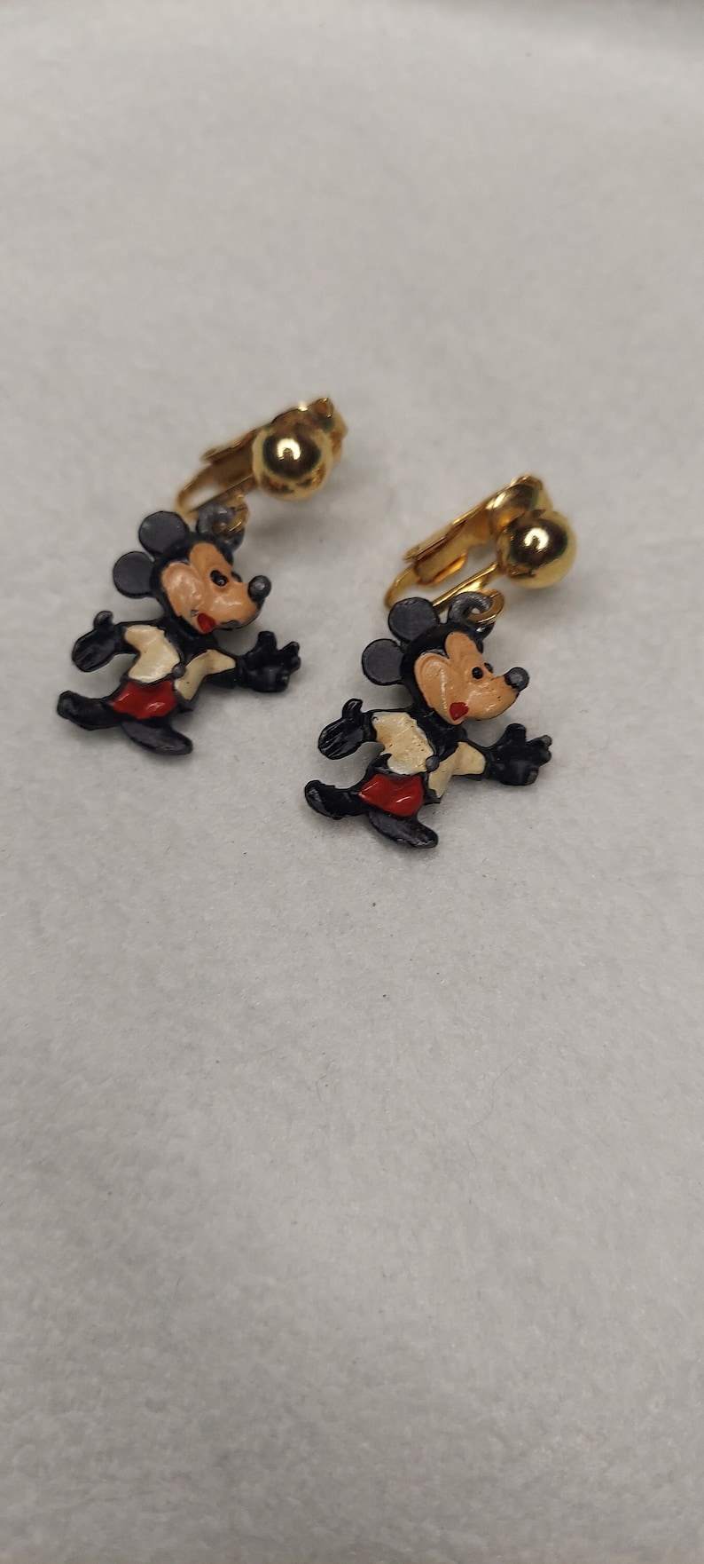 May include: A pair of clip-on earrings featuring a running Mickey Mouse design. The earrings have a gold-tone finish with black, red, and white enamel details. The earrings are a classic Disney collectible.