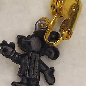 May include: A vintage clip-on earring featuring a black cartoon character with a gold-tone clip. The character is wearing a vest and hat. The earring is set against a white background.