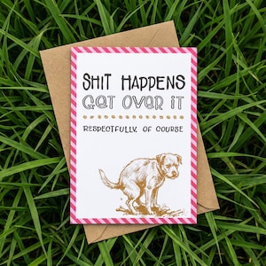 May include: A greeting card with the text "Shit Happens Get Over It Respectfully Of Course." The card has a pink and white striped border and a brown dog illustration. It is placed on a brown envelope, set against a backdrop of green grass.
