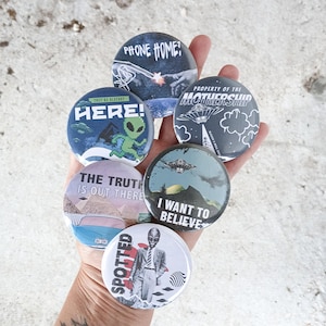 May include: A collection of six round buttons with alien-themed designs. The buttons feature phrases like "PHONE HOME?" and "I WANT TO BELIEVE," along with images of UFOs and aliens. The color palette includes blues, greens, and whites.