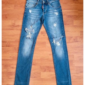Puede incluir: Pantalones vaqueros de mezclilla azul con un acabado desgastado que presenta múltiples roturas y zonas deshilachadas en los muslos y las rodillas. Los pantalones incluyen cierre de botones, cinco bolsillos y una etiqueta roja en la cintura.