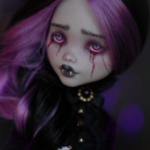 May include: A gothic-style doll with black hair and purple highlights. The doll has pale skin, dark eye makeup, and tears of blood. It wears a black outfit with a decorative collar and has fangs. The doll's expression is somber.