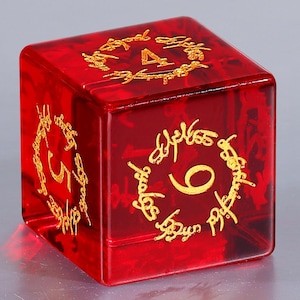 May include: A translucent red six-sided die featuring gold-colored numbers four, five, and six on its visible faces. Each number is encircled by intricate, decorative gold script lettering that mimics an ancient, flowing calligraphic style.