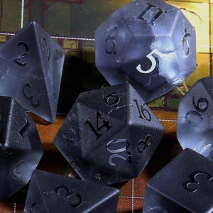 May include: Set of seven translucent, dark gray polyhedral dice for tabletop gaming. Each die features white numbering and a unique geometric shape, including a d4, d6, d8, d10, d12, d20, and a percentile die.