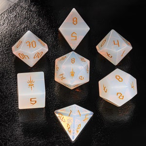 May include: Set of seven opaque white dice with gold numbers and symbols. The dice are a d4, d6, d8, d10, d%, d12, and d20.