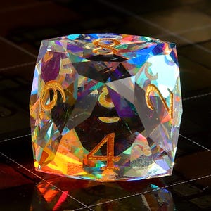 May include: A clear iridescent crystal dice with gold numbers 1, 2, 3, 4, 5, and 6. The dice is face down with the number 4 showing.