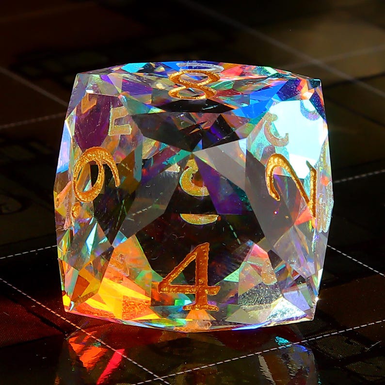 May include: A clear iridescent crystal dice with gold numbers 1, 2, 3, 4, 5, and 6. The dice is face down with the number 4 showing.