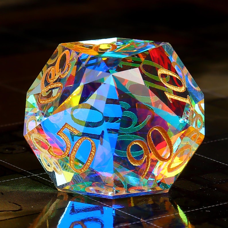 May include: A multi-faceted iridescent crystal dice with gold numbers. The dice is a 10-sided die with numbers 0, 10, 20, 30, 40, 50, 60, 70, 80, and 90.