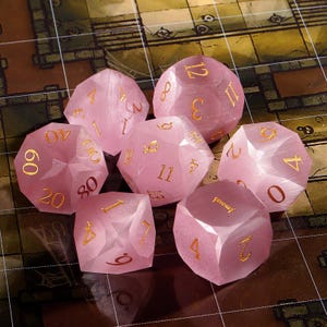 May include: A set of seven polyhedral dice in a translucent light pink color with a cat's eye effect. Each die features engraved numbers filled with gold-colored ink, including a d20, d12, d10, d8, d6, d4, and a percentile die with tens digits.