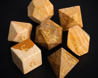 May include: Set of seven stone dice in various shapes and sizes. The dice are a light brown color with gold numbers. The dice are on a black surface.