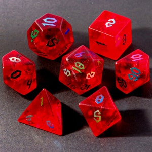 Op de afbeelding: Set van zeven rode dobbelstenen met regenboogkleurige, iriserende nummers. De dobbelstenen zijn gemaakt van helder plastic met een rode tint en hebben een gespikkeld uiterlijk. De nummers op de dobbelstenen zijn in zwart lettertype met een regenboogkleurige, iriserende glans.