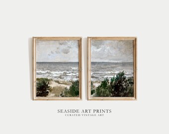 May include: Two framed paintings of a seascape with a cloudy sky, blue water, and green bushes in the foreground. The paintings are in wooden frames with a light brown finish. The text "SEASIDE ART PRINTS CURATED VINTAGE ART" is below the paintings.