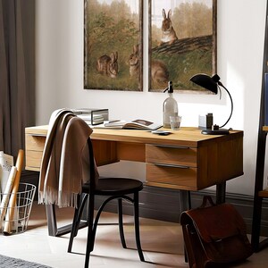 Op de afbeelding: Een houten bureau met zwarte metalen poten, twee laden en een zwarte stoel. Twee ingelijste prints van konijnen in een bossetting hangen aan de muur boven het bureau. Een bruine leren tas ligt op de vloer naast het bureau.