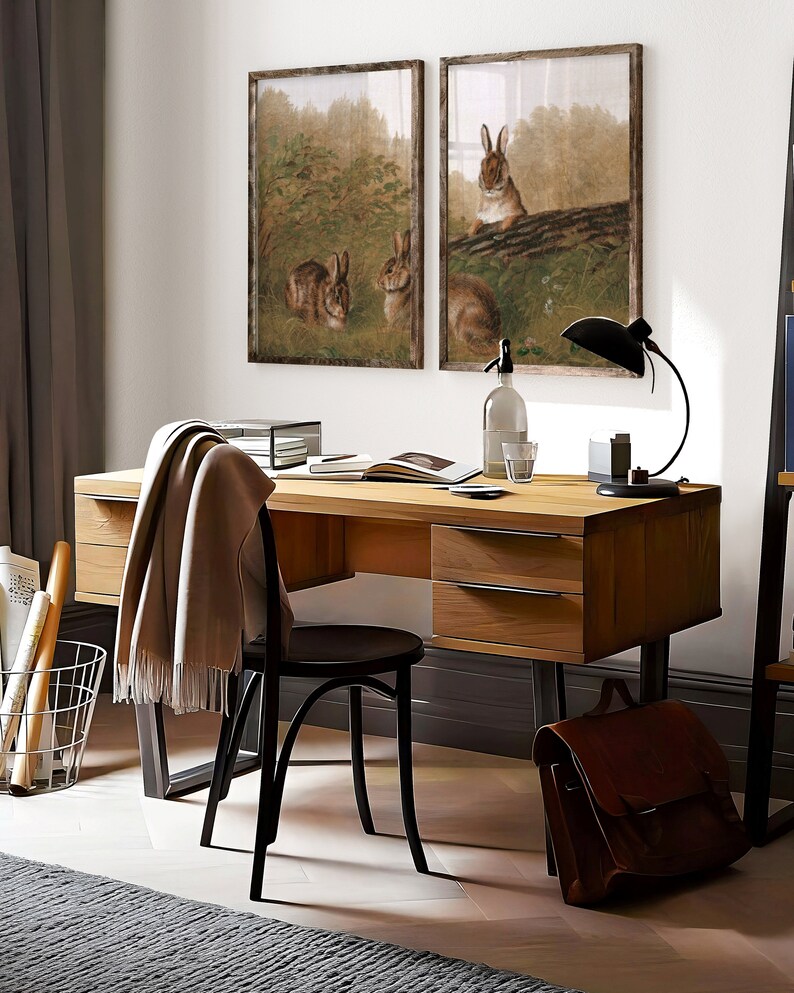 Op de afbeelding: Een houten bureau met zwarte metalen poten, twee laden en een zwarte stoel. Twee ingelijste prints van konijnen in een bossetting hangen aan de muur boven het bureau. Een bruine leren tas ligt op de vloer naast het bureau.