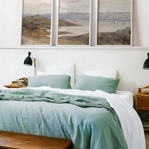 May include: A bedroom with a bed with a white and green linen duvet cover and two green pillows. There are three framed landscape paintings above the bed on a white wall with white wainscoting. There is a wooden nightstand on the right side of the bed with a lamp and a small wooden bench at the foot of the bed.
