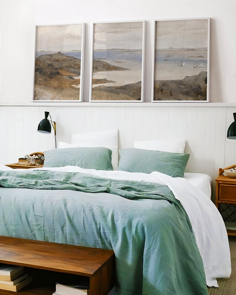 May include: A bedroom with a bed with a white and green linen duvet cover and two green pillows. There are three framed landscape paintings above the bed on a white wall with white wainscoting. There is a wooden nightstand on the right side of the bed with a lamp and a small wooden bench at the foot of the bed.