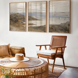 May include: Three framed landscape paintings of a seascape with a rocky shoreline and a body of water in the distance. The paintings are hung on a white wall above a wicker coffee table and a wooden chair with a white cushion.