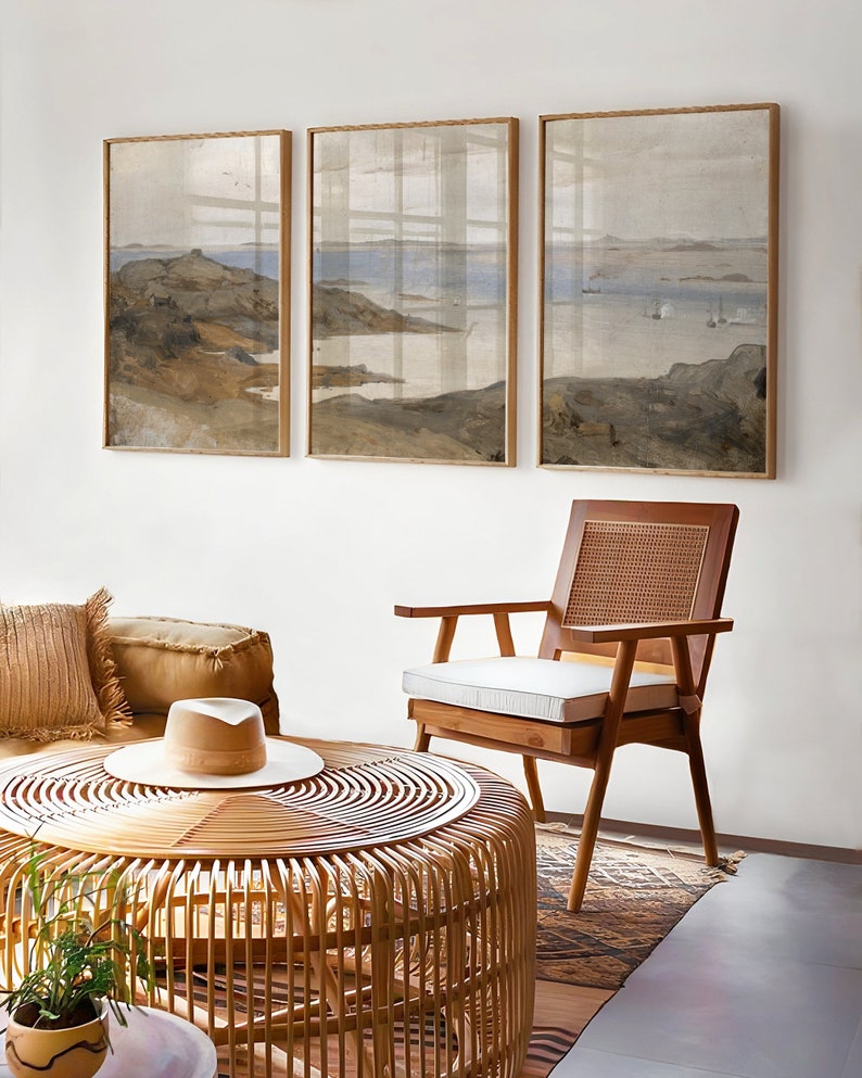 May include: Three framed landscape paintings of a seascape with a rocky shoreline and a body of water in the distance. The paintings are hung on a white wall above a wicker coffee table and a wooden chair with a white cushion.