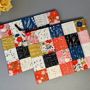 May include: Two patchwork zipper pouches with floral and geometric patterns in red, pink, blue, black, and gold. The pouches have a black zipper pull and a small fabric loop. The background is a light grey.