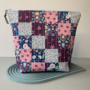 May include: A quilted zippered pouch with a patchwork design featuring floral patterns in shades of pink, blue, and purple. The pouch is made of fabric and has a zipper closure.