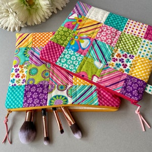 May include: Two colourful patchwork zippered pouches with a yellow zipper and pink tassel. The pouches are filled with makeup brushes.
