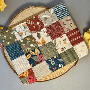 May include: A patchwork zipped pouch featuring floral and geometric patterns in green, red, and beige. The pouch is placed on a wooden slice and has a yellow zip pull.